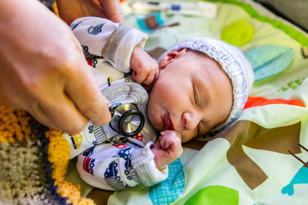 A newborn baby wearing a knit hat and patterned onesie is examined with a stethoscope, lying peacefully on a colorful blanket.